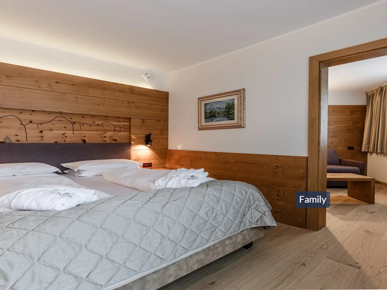 Komfortowy pokój hotelowy z dużym łóżkiem, drewnianym zagłówkiem i szlafrokami. Widok na salon z sofą, idealny dla rodzin.