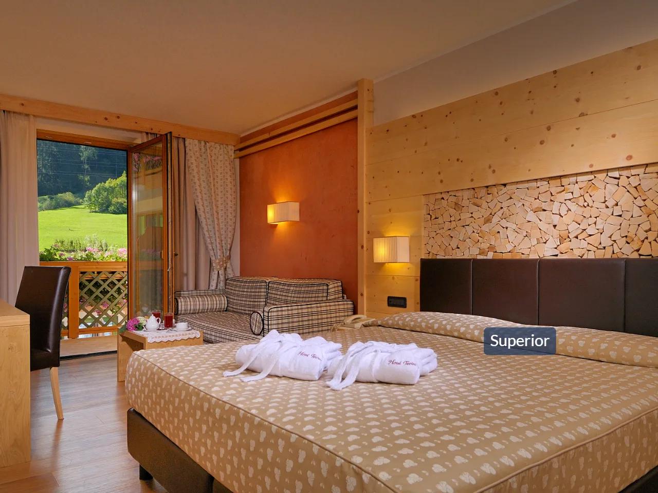 Pokój hotelowy Superior w TEVINI, Val di Sole we Włoszech z dużym łóżkiem, balkonem i widokiem na zieloną dolinę.