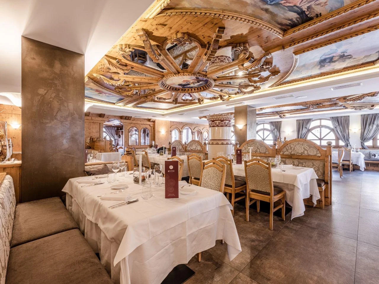 Restauracja hotelowa z eleganckimi stolikami, regionalnym wystrojem oraz bogato zdobionym drewnianym sufitem.