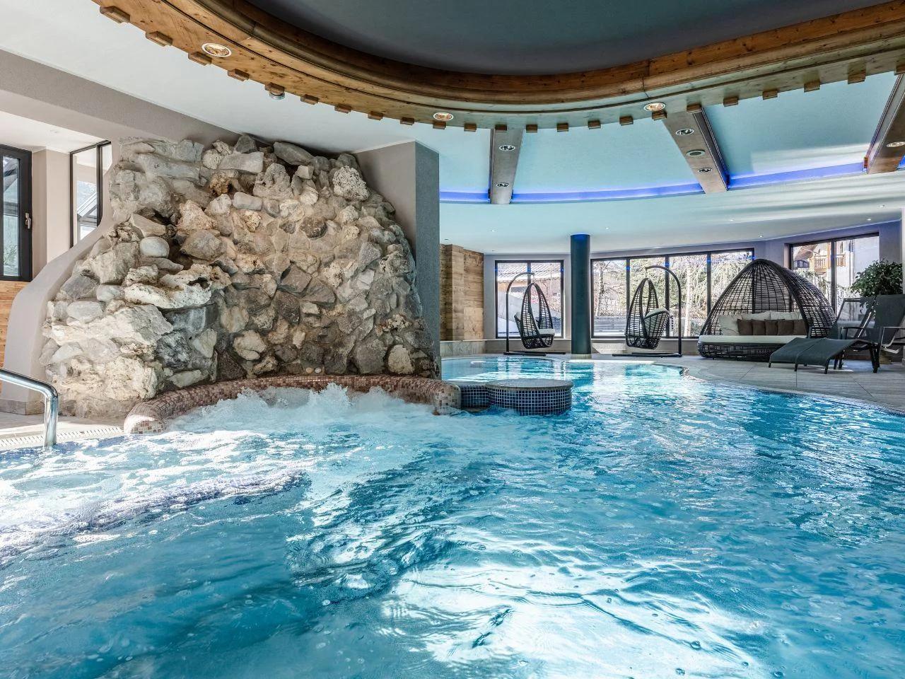 Luksusowy basen wewnętrzny w hotelu Tevini Val di Sole we Włoszech, z błękitną wodą, strefą jacuzzi, drewnianym sufitem i oknami.