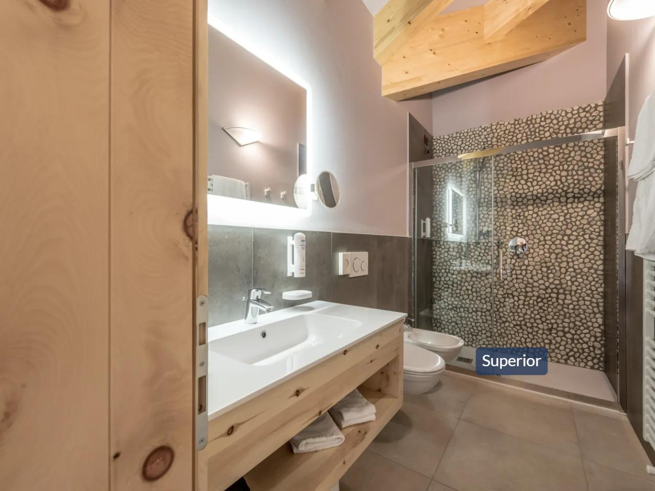 Nowoczesna łazienka hotelu Tevini w Val di Sole, Włochy. Drewniana szafka, podświetlane lustro i prysznic z kamiennym motywem.