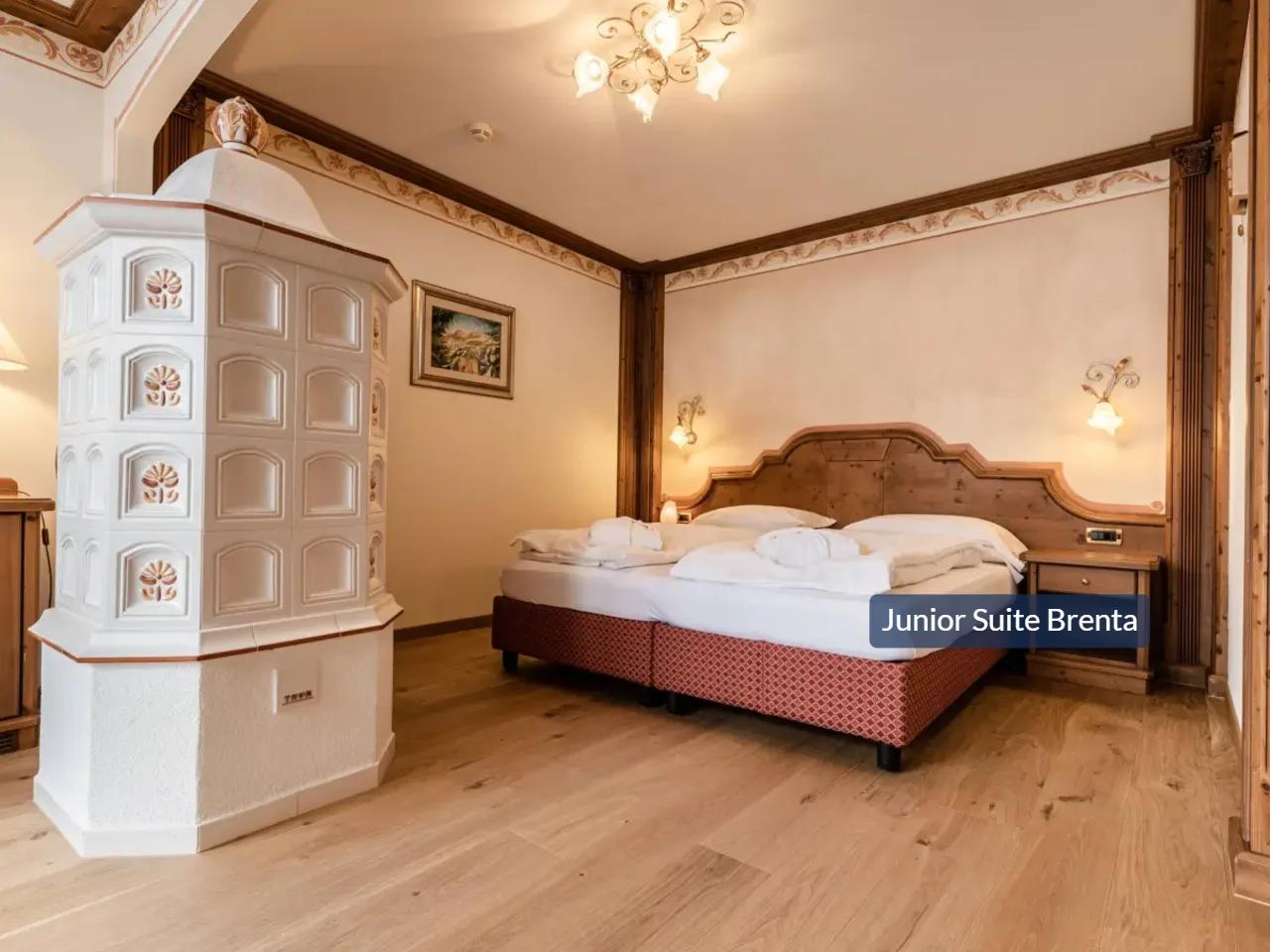 Apartament Junior Suite Brenta w hotelu Tevini, Val di Sole, Włochy. Przytulna sypialnia z łóżkiem i piecem kaflowym.