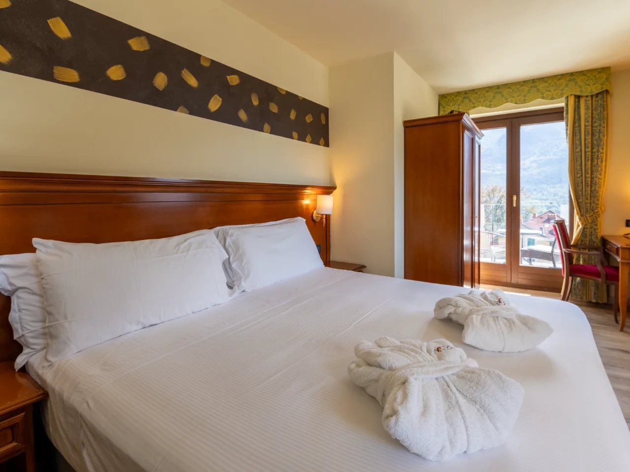 Elegancki pokój hotelowy Au Soleil w Aosta, Włochy, z dużym łóżkiem, szlafrokami, balkonem i drewnianymi meblami.