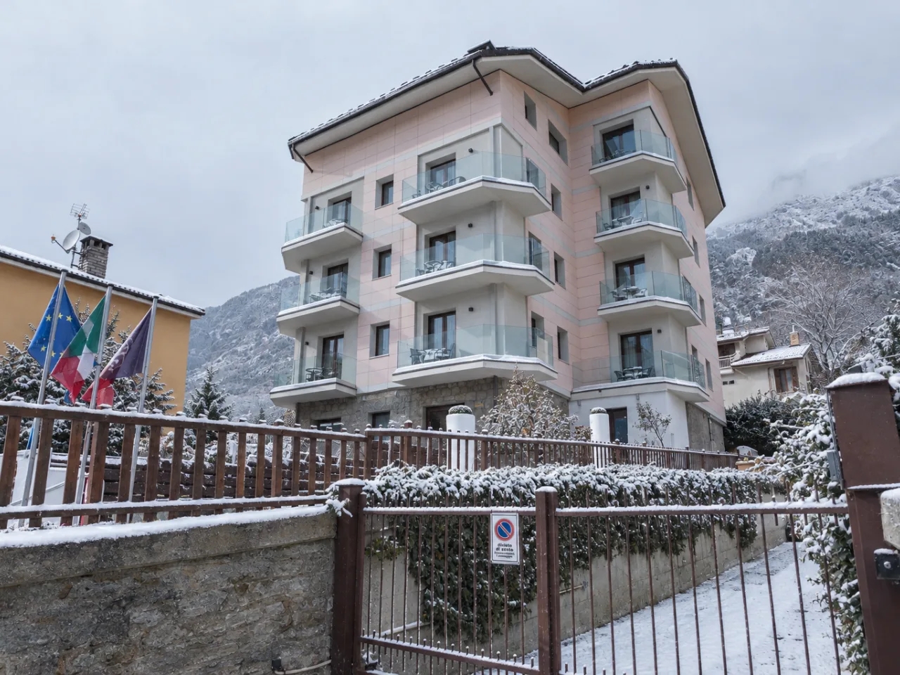 Zimowy widok na Hotel Au Soleil w Aosta, Włochy, z ośnieżonymi balkonami, ogrodzeniem i drzewami, pod zachmurzonym niebem.