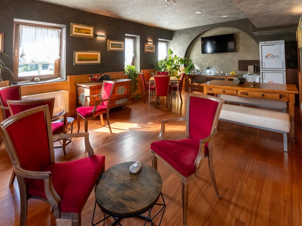 Wnętrze hotelu Au Soleil w Aosta: elegancka jadalnia lub salon z czerwonymi fotelami, drewnianymi meblami i dziełami sztuki.