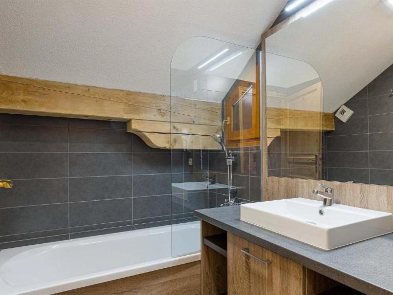 Nowoczesna łazienka w apartamencie La Cascade Alpe D'Huez: wanna, prysznic, umywalka, szare płytki i drewniane belki.