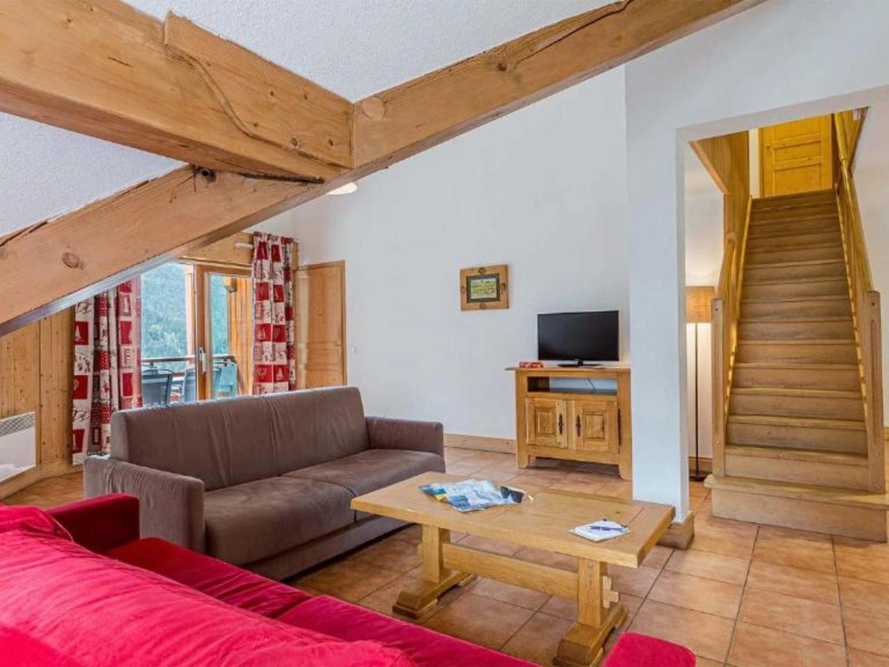 Przytulny salon w stylu górskiego chalet z drewnianymi belkami, czerwoną sofą, telewizorem i schodami, obiekt La Cascade, Alpe d'Huez.