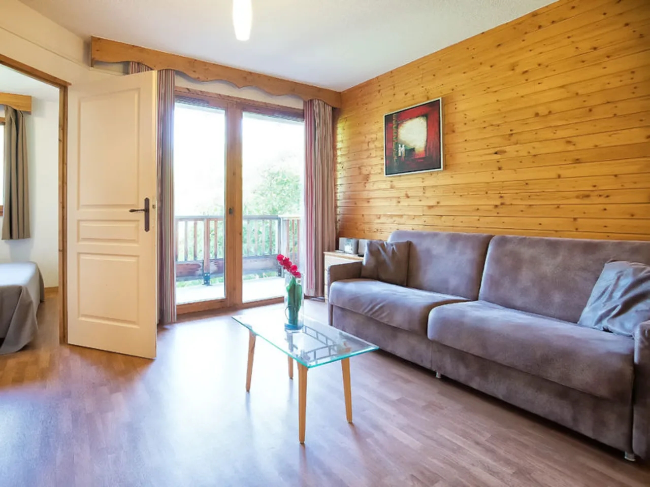 Przytulny salon z szarą sofą, drewnianą ścianą i szklanym stolikiem. Widok na balkon, sypialnia obok.