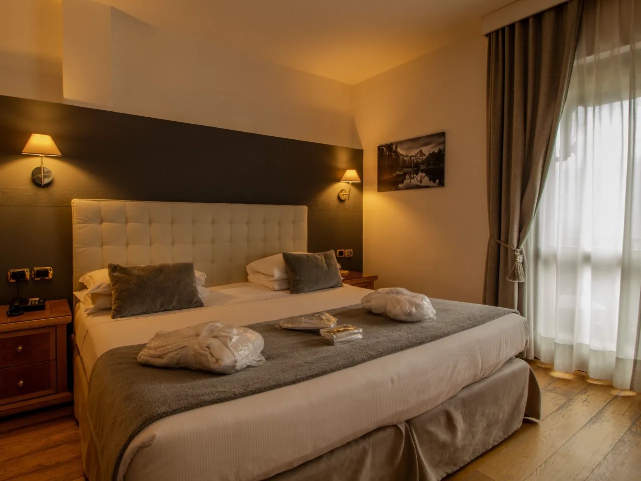 Luksusowy pokój hotelowy z dużym łóżkiem, białym zagłówkiem, szarą narzutą, eleganckimi lampami i drewnianą podłogą.