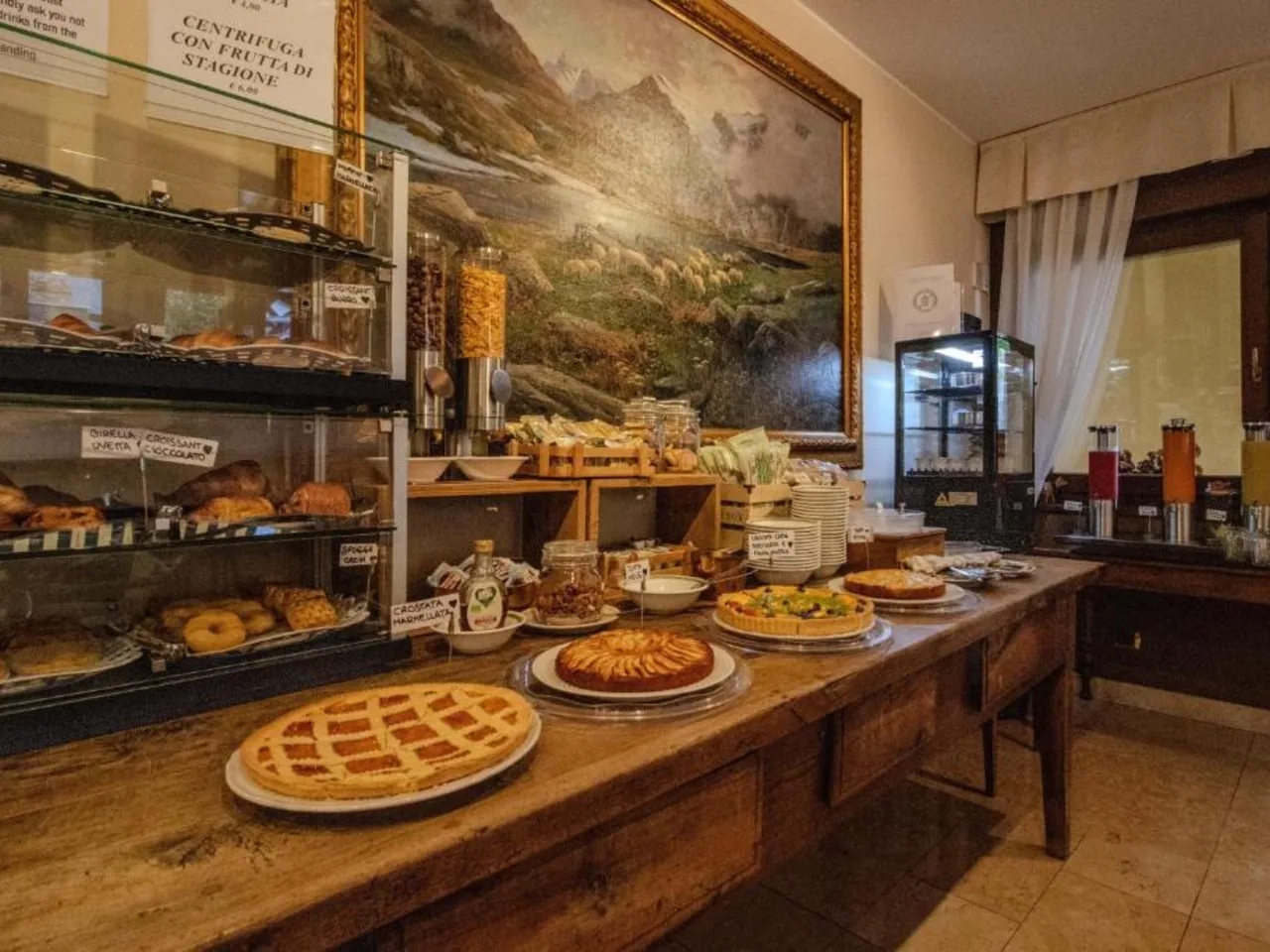 Hotelowy bufet śniadaniowy: ciasta, świeże soki, pieczywo i płatki. W tle malowniczy obraz górskiego krajobrazu.