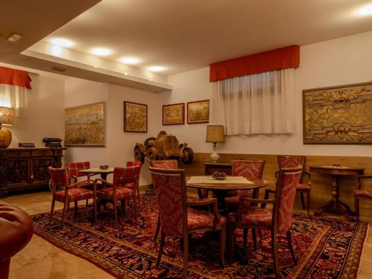 Przytulny salon w stylu klasycznym z okrągłymi stołami, czerwonymi krzesłami, orientalnym dywanem i unikatowymi dekoracjami ściennymi.