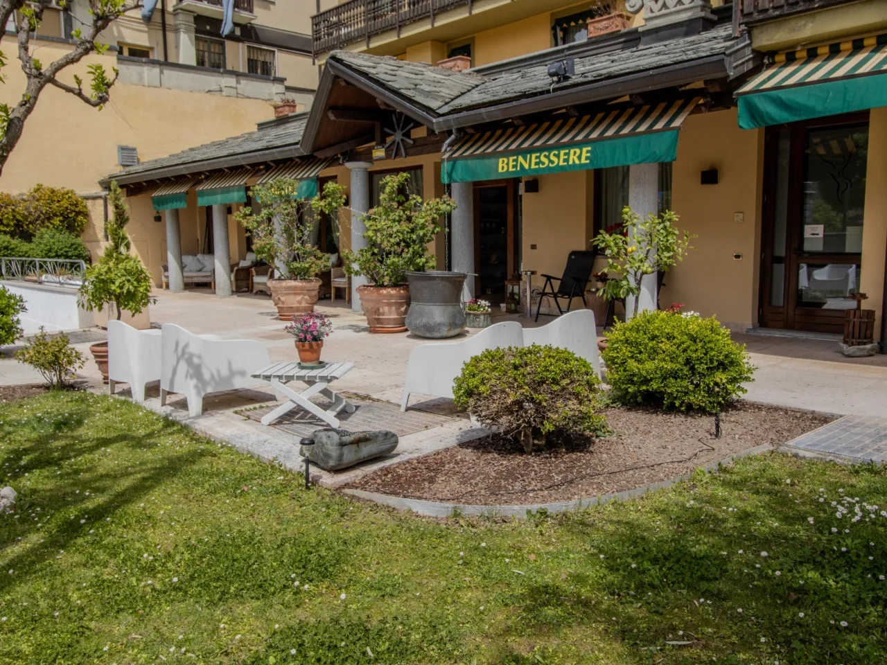 Taras hotelowy z ogrodem i miejscami do siedzenia. W tle budynek z zadaszeniem i napisem 'BENESSERE'. Idealne miejsce na relaks.