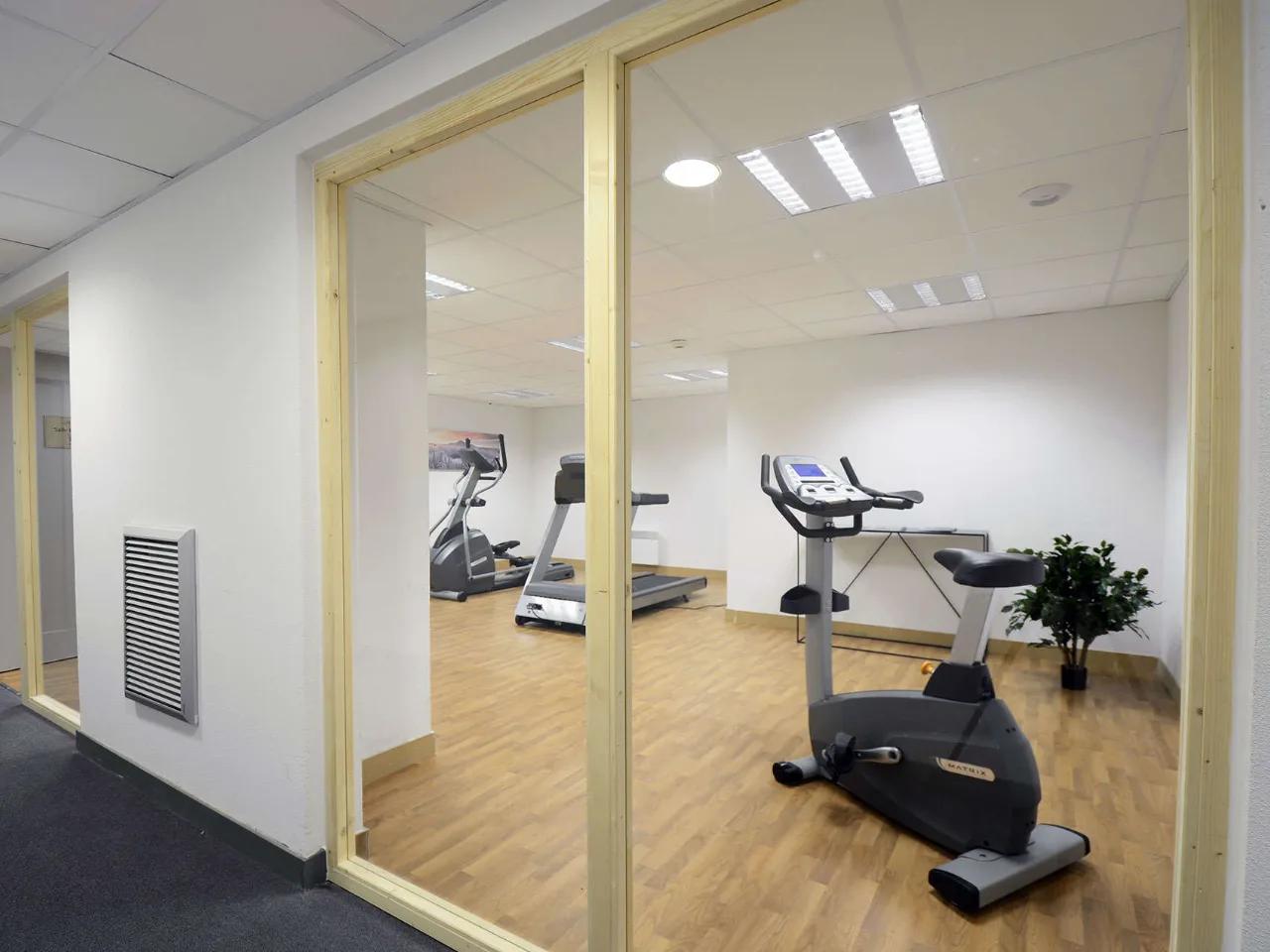 Nowoczesna siłownia biurowa ze sprzętem fitness: rower stacjonarny, bieżnia, orbitrek, widoczna przez szklaną ścianę.