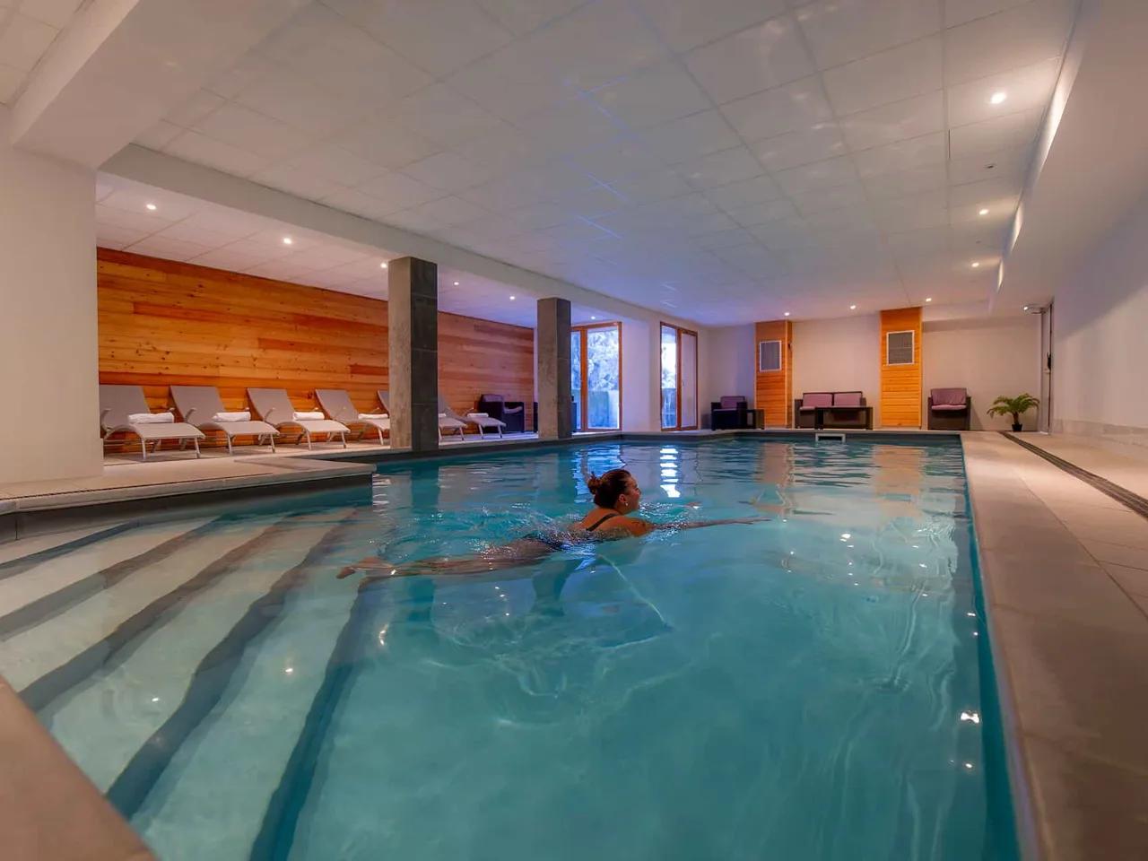 Kobieta pływająca w nowoczesnym krytym basenie hotelowym. W tle drewniane ściany, leżaki i duże okna, idealne do relaksu.